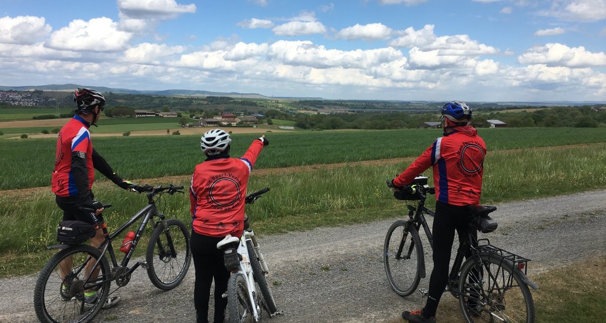 Drei Radfahrer in roten Trikots stehen auf einem Feldweg und schauen in die weite Landschaft. Einer zeigt in die Ferne. Der Himmel ist bewölkt., © Land der 1000 Hügel - Kraichgau-Stromberg Drei Radfahrer in roten Trikots stehen auf einem Feldweg und schauen in die weite Landschaft. Einer zeigt in die Ferne. Der Himmel ist bewölkt., © Land der 1000 Hügel - Kraichgau-Stromberg