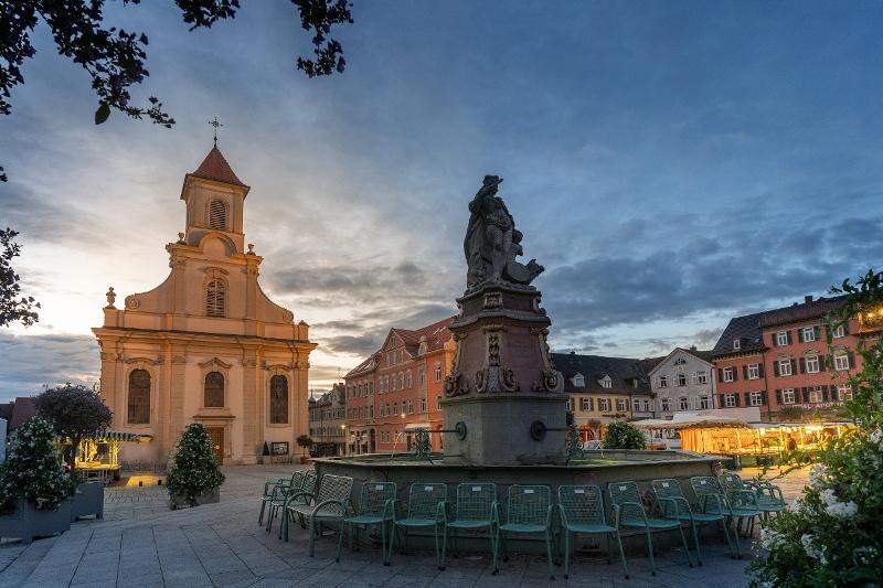 Abendstimmung auf einem Platz in Ludwigsburg. Eine Kirche und ein Brunnen sind im Vordergrund, umgeben von historischen Gebäuden und Stühlen., © © Martina Denker