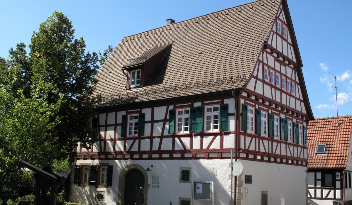 Fachwerkhaus mit gr&uuml;n-wei&szlig;en Fensterl&auml;den und Satteldach, umgeben von B&auml;umen und blauem Himmel., &copy; Natur.Nah. Sch&ouml;nbuch & Heckeng&auml;u