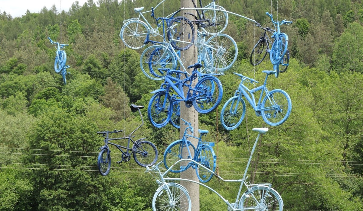 Blau gestrichene Fahrr&auml;der h&auml;ngen als Kunstinstallation an einem Pfosten in einer gr&uuml;nen, bewaldeten Landschaft., &copy; Natur.Nah. Sch&ouml;nbuch & Heckeng&auml;u