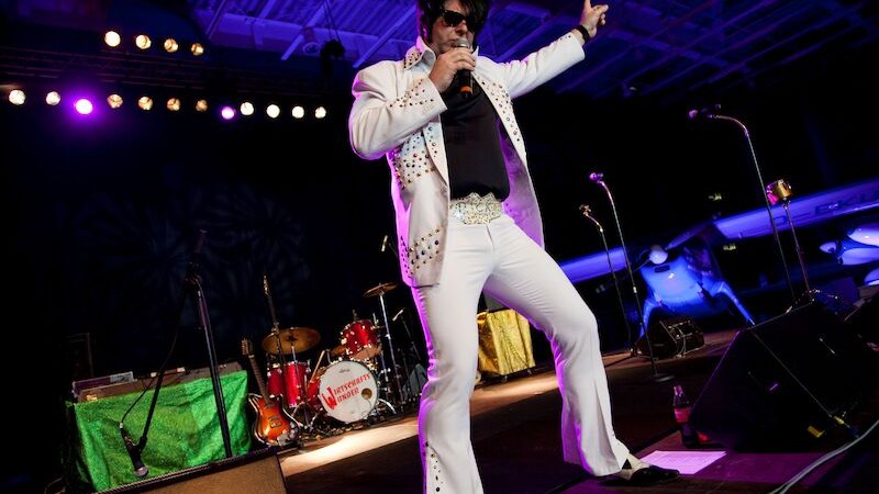 Ein Elvis-Imitator in einem wei&szlig;en Anzug performt auf einer B&uuml;hne. Im Hintergrund sind Instrumente und bunte Lichter zu sehen., &copy; Touristik Bad Wildbad GmbH