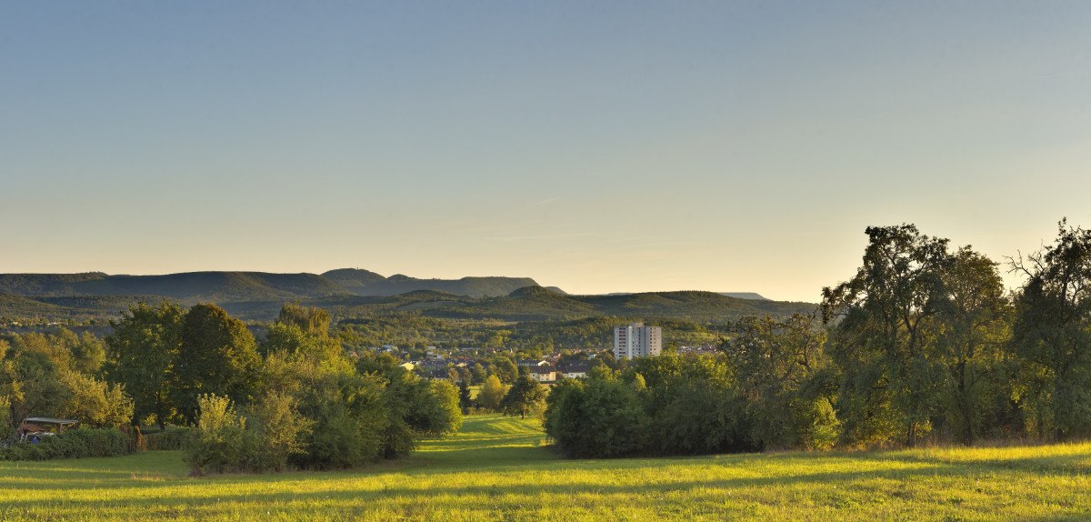 Grüne Wiesen und Bäume im Vordergrund, dahinter die Stadt Betzingen. Im Hintergrund erheben sich die Hügel Käpfle und Rossberg unter klarem Himmel., © Mythos Schwäbische Alb Grüne Wiesen und Bäume im Vordergrund, dahinter die Stadt Betzingen. Im Hintergrund erheben sich die Hügel Käpfle und Rossberg unter klarem Himmel., © Mythos Schwäbische Alb