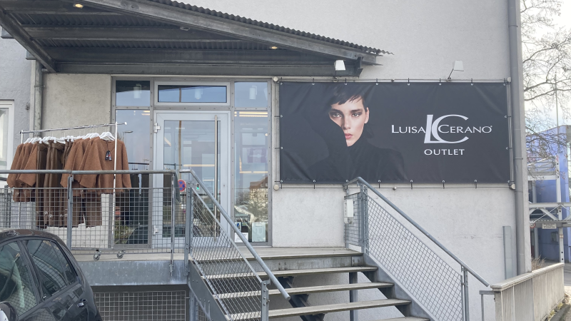 Eingang eines Luisa Cerano Outlets mit Treppe, Werbebanner und Kleidung auf einem St&auml;nder. Ein Auto parkt daneben, B&uuml;sche im Vordergrund., &copy; L. Besmer