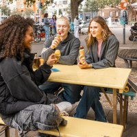 Drei Personen sitzen an einem gelben Tisch im Freien und essen Eis. Im Hintergrund sind B&auml;ume, Fahrr&auml;der und ein Caf&eacute; zu sehen., &copy; Stuttgart Marketing GmbH, Sarah Schmid