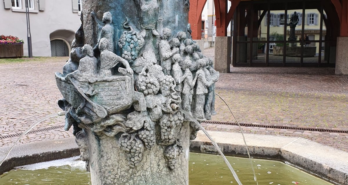 Brunnen mit detaillierten Skulpturen und Wasserspeiern, umgeben von Fachwerkhäusern und einem gepflasterten Platz., © Kulturamt Stadt Plochingen Brunnen mit detaillierten Skulpturen und Wasserspeiern, umgeben von Fachwerkhäusern und einem gepflasterten Platz., © Kulturamt Stadt Plochingen