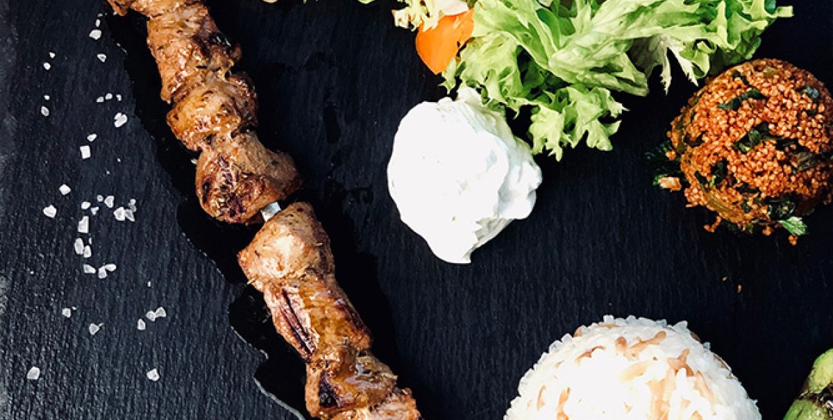 Gegrillter Fleischspieß mit Reis, Salat, Bohnen, gegrilltem Gemüse und weißer Soße auf schwarzem Untergrund angerichtet., © Bady's