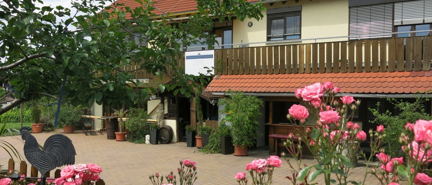 Ein Weinhof mit Holzbalkon, roten Dachziegeln und blühenden Rosen im Vordergrund. Ein Schild mit der Aufschrift 'Weinhof Württemberg Helmut Zaiß' ist sichtbar., © Helmut Zaiß Weingut & Weingastronomie