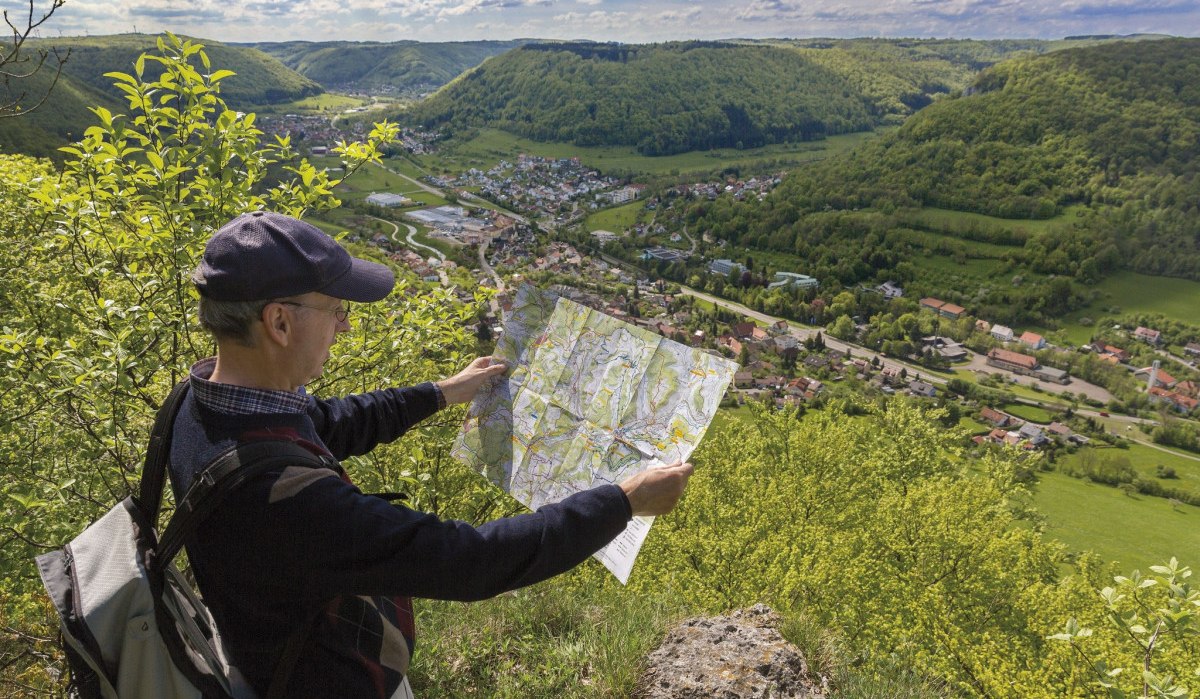 Ein Wanderer betrachtet eine Karte, während er auf ein grünes Tal mit einem Dorf und bewaldeten Hügeln blickt., © Landkreis Göppingen Ein Wanderer betrachtet eine Karte, während er auf ein grünes Tal mit einem Dorf und bewaldeten Hügeln blickt., © Landkreis Göppingen