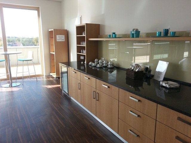 Moderne Teeküche mit Holzschränken, Geschirr und Kaffeemaschine. Heller Raum mit großem Fenster und Tisch mit Stühlen im Hintergrund., © TOMAS Moderne Teeküche mit Holzschränken, Geschirr und Kaffeemaschine. Heller Raum mit großem Fenster und Tisch mit Stühlen im Hintergrund., © TOMAS