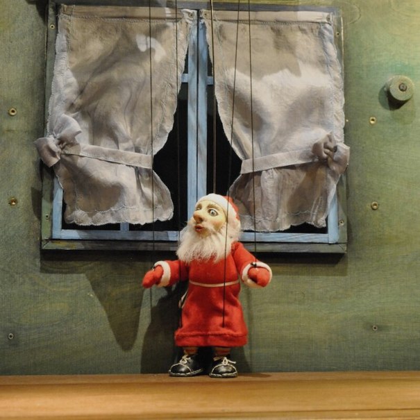 Eine Marionette in rotem Kostüm steht vor einem Fenster mit weißen Vorhängen. Die Szene wirkt wie aus einem Puppentheater., © Mauerwerk GmbH