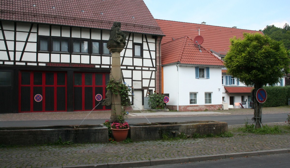 Fachwerkhaus mit rotem Dach, Feuerwehr-Gerätehaus. Im Vordergrund ein Brunnen mit Blumen. Parkverbotsschilder und ein Baum sind sichtbar., © Natur.Nah. Schönbuch & Heckengäu