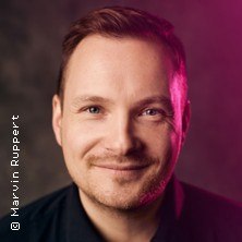 Ein Mann lächelt in die Kamera, während ein pinker Lichteffekt sein Gesicht beleuchtet. Der Hintergrund ist dunkel gehalten., © links im Bild Ein Mann lächelt in die Kamera, während ein pinker Lichteffekt sein Gesicht beleuchtet. Der Hintergrund ist dunkel gehalten., © links im Bild