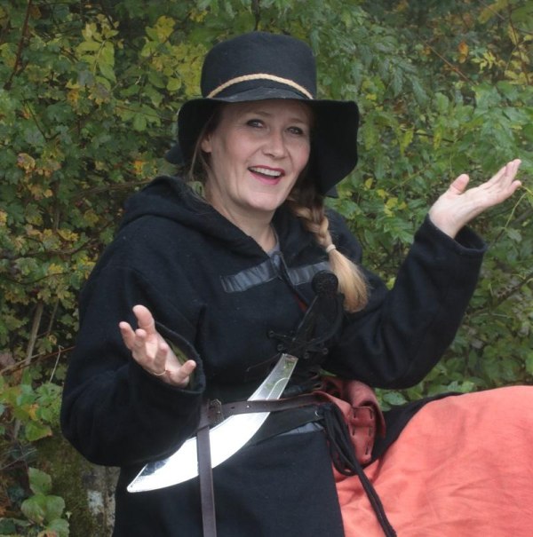 Eine Frau in mittelalterlicher Kleidung mit einem schwarzen Hut und einem Schwert posiert l&auml;chelnd im Wald., &copy; Copyright: Stefanie Keller