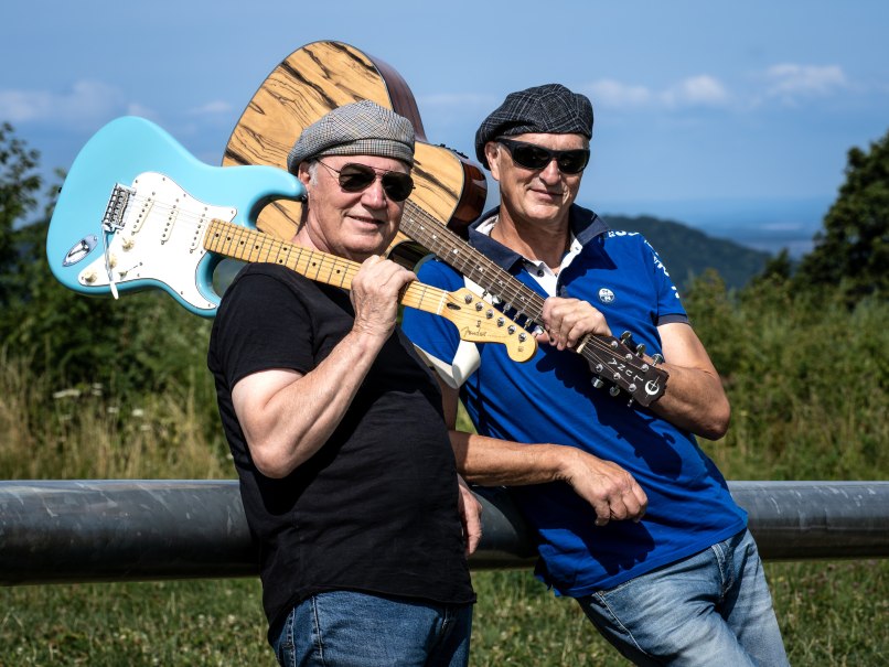Zwei M&auml;nner mit Gitarren posieren l&auml;ssig im Freien. Sie tragen Sonnenbrillen und M&uuml;tzen, im Hintergrund ist eine h&uuml;gelige Landschaft zu sehen., &copy; (c) G&uuml;nter Orban