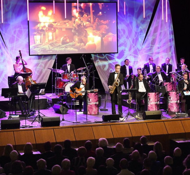 Eine Bigband spielt auf einer Bühne, im Hintergrund weihnachtliche Projektionen. Musiker in Anzügen, Publikum im Vordergrund., © Stadt Leinfelden-Echterdingen, Kulturamt Eine Bigband spielt auf einer Bühne, im Hintergrund weihnachtliche Projektionen. Musiker in Anzügen, Publikum im Vordergrund., © Stadt Leinfelden-Echterdingen, Kulturamt