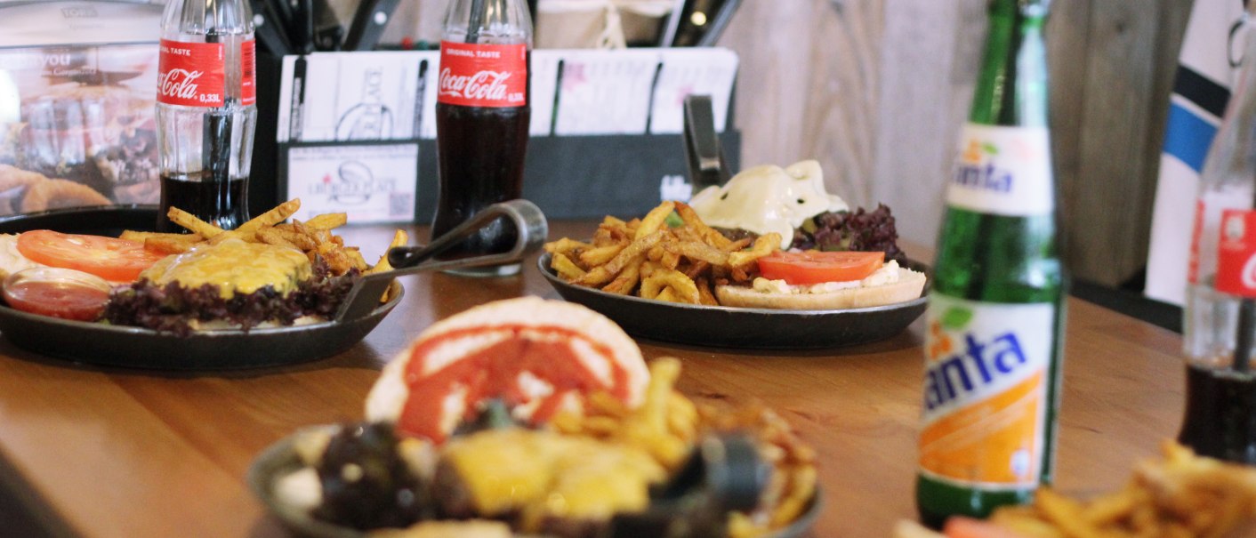 Burger mit Pommes und Getränken wie Cola und Fanta auf einem Holztisch in einem Restaurant., © SMG