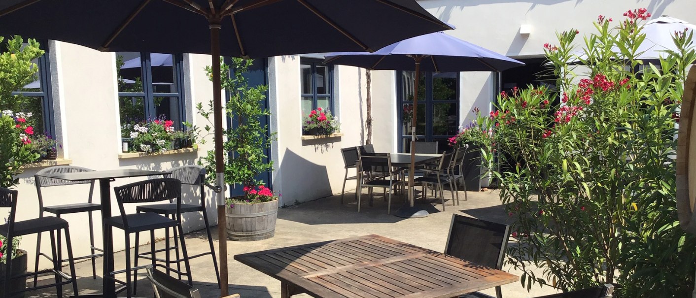 Sonnige Terrasse eines Weinguts mit Holztischen, Stühlen und blauen Sonnenschirmen. Blumen und Pflanzen schmücken den Außenbereich., © Weingut Wöhrwag