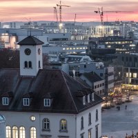 Stadtansicht bei Sonnenuntergang: Ein Kirchturm im Vordergrund, moderne Gebäude und Baukräne im Hintergrund, beleuchtete Fenster und ein farbenfroher Himmel., © Stadt Sindelfingen