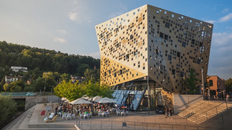 Modernes Gebäude mit goldener Fassade, viele Fenster. Menschen sitzen unter Sonnenschirmen. Umgeben von Bäumen und Treppen., © Remstal Tourismus e.V. I Architekt: isin Architekten