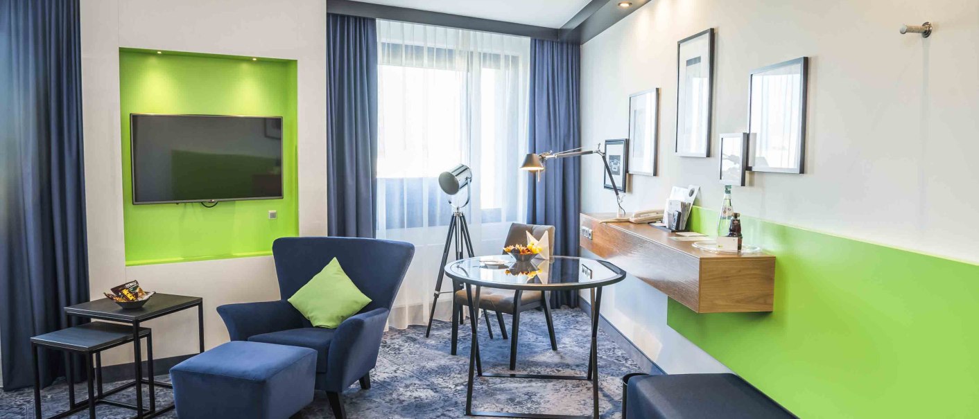 Modernes Hotelzimmer mit blauem Sessel, grünem Akzent, Flachbildfernseher, Schreibtisch und stilvoller Beleuchtung., © Chandler Photography Modernes Hotelzimmer mit blauem Sessel, grünem Akzent, Flachbildfernseher, Schreibtisch und stilvoller Beleuchtung., © Chandler Photography