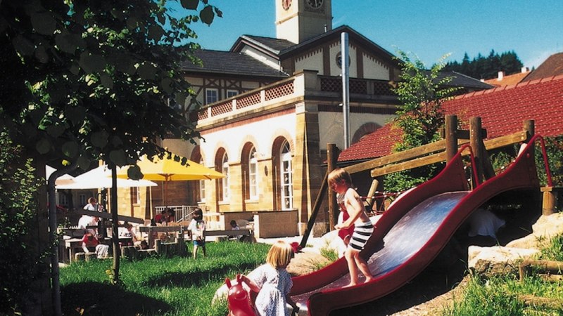 Kinder spielen auf einer Rutsche vor einem historischen Gebäude mit Turm und Sonnenschirmen im Garten., © Stuttgart-Marketing GmbH Kinder spielen auf einer Rutsche vor einem historischen Gebäude mit Turm und Sonnenschirmen im Garten., © Stuttgart-Marketing GmbH