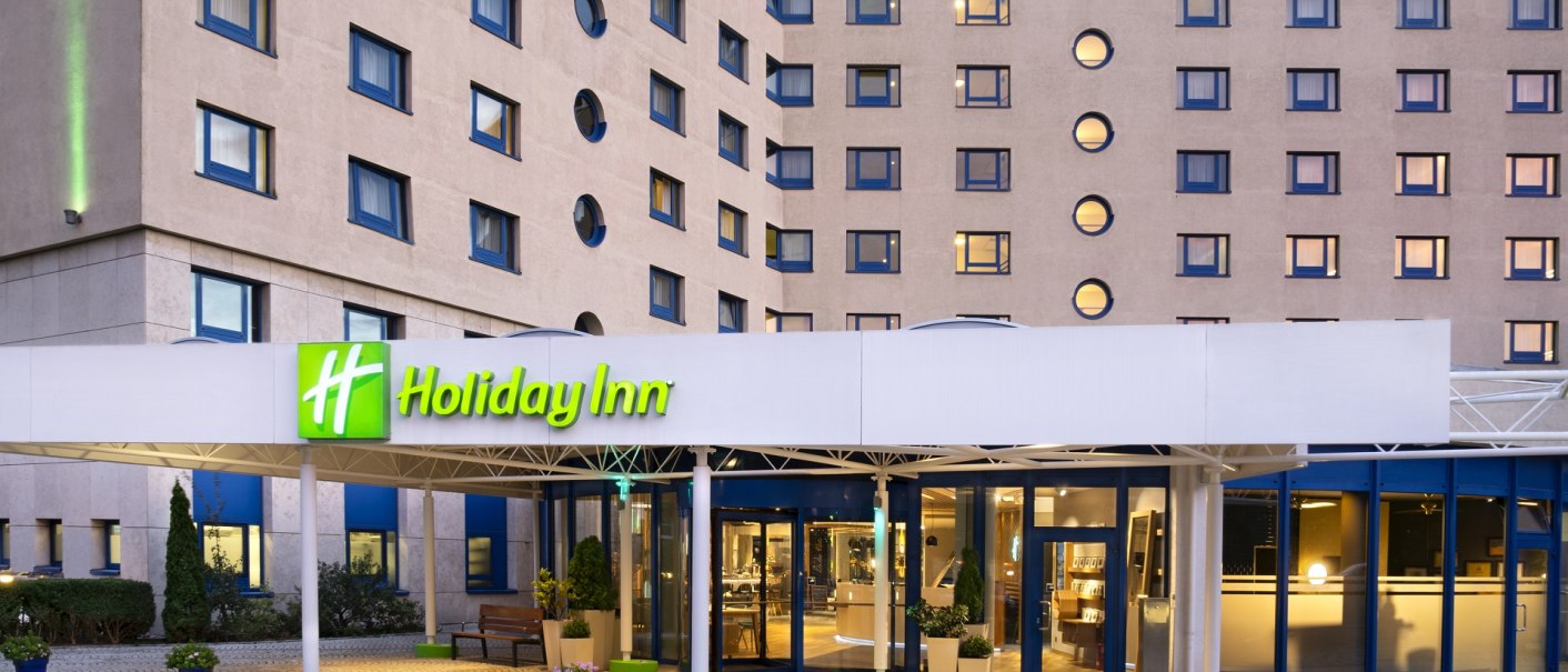 Außenansicht eines Holiday Inn Hotels mit beleuchtetem Eingang, runden und rechteckigen Fenstern. Gepflasterter Vorplatz mit Pflanzen., © Holiday Inn Stuttgart Außenansicht eines Holiday Inn Hotels mit beleuchtetem Eingang, runden und rechteckigen Fenstern. Gepflasterter Vorplatz mit Pflanzen., © Holiday Inn Stuttgart