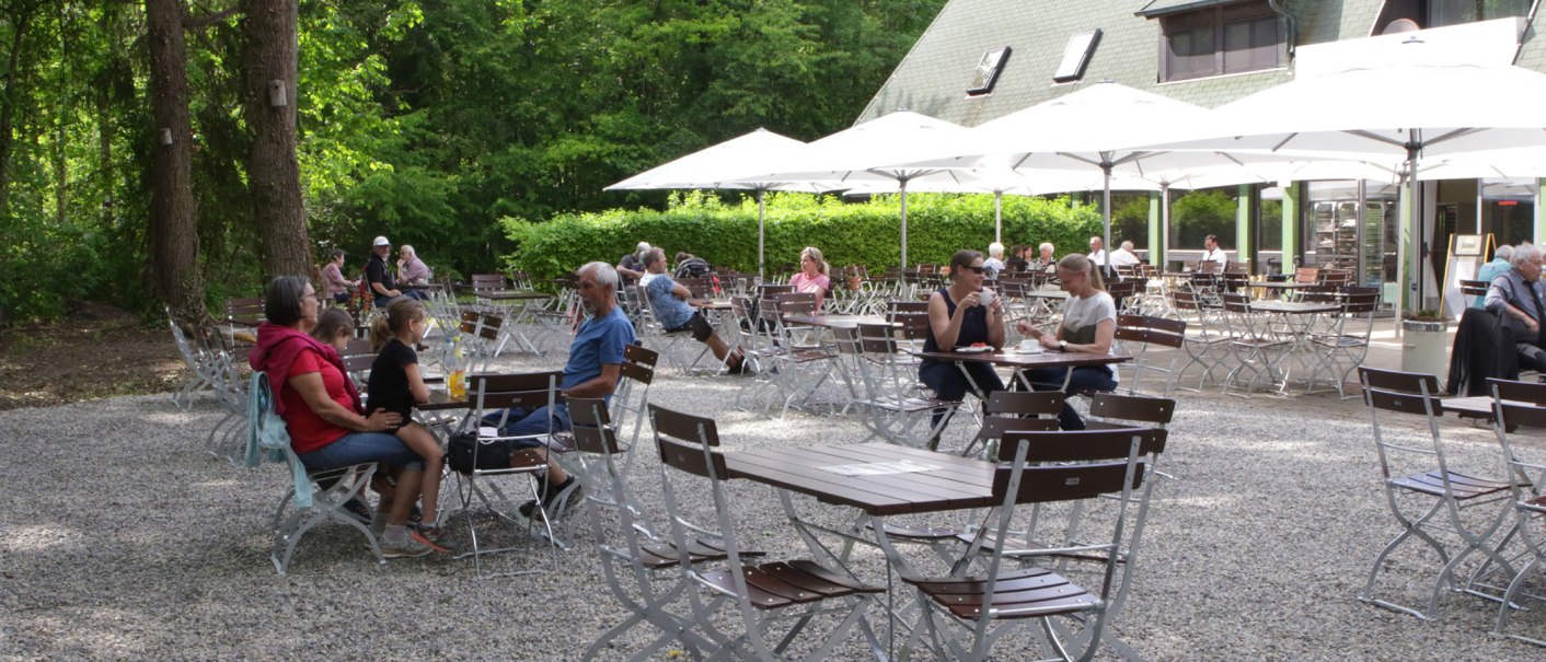 Menschen sitzen im Außenbereich eines Cafés mit weißen Sonnenschirmen, umgeben von Bäumen und grüner Vegetation., © Gabriel Holom