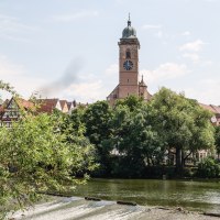 Ein Kirchturm erhebt sich &uuml;ber den Neckar, umgeben von B&auml;umen und Fachwerkh&auml;usern. Im Vordergrund flie&szlig;t der Fluss &uuml;ber ein Wehr., &copy; Belsers Hotel