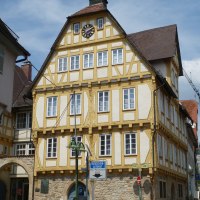 Ein historisches Fachwerkhaus mit gelben Balken, Uhr und Glockenturm. Es steht an einer gepflasterten Straße mit Parkscheinautomat und Blumenkübel. Ein historisches Fachwerkhaus mit gelben Balken, Uhr und Glockenturm. Es steht an einer gepflasterten Straße mit Parkscheinautomat und Blumenkübel.