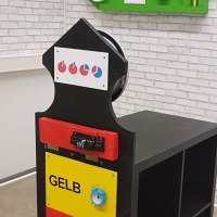 Escape-Room-Puzzle mit farbigen Schubladen und Symbolen. An der Wand eine grüne Dekoration mit Uhr und Rätseln., © Flucht-Raum