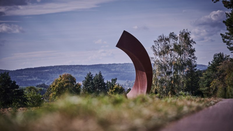 Große, gebogene Skulptur in einer Landschaft mit Bäumen und Hügeln im Hintergrund unter blauem Himmel., © Natur.Nah. Schönbuch & Heckengäu Große, gebogene Skulptur in einer Landschaft mit Bäumen und Hügeln im Hintergrund unter blauem Himmel., © Natur.Nah. Schönbuch & Heckengäu