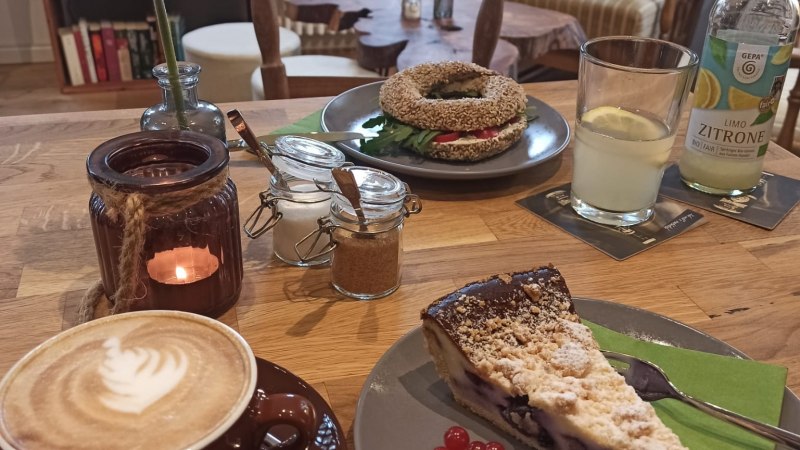 Gem&uuml;tliches Caf&eacute; mit Cappuccino, Kuchen und Bagel auf einem Holztisch. Im Hintergrund B&uuml;cherregale und bequeme Sessel., &copy; KaffeehausMaLe