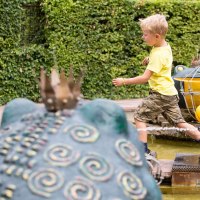 Ein Junge in gelbem T-Shirt und Tarnshorts läuft über Steine in einem Märchengarten. Im Hintergrund sind Froschskulpturen und Wasserfontänen zu sehen., © Tourismus & Events Ludwigsburg
