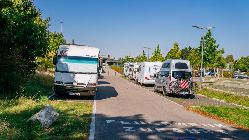 Wohnmobilstellplatz in Fellbach mit geparkten Wohnmobilen und Autos, umgeben von Bäumen und klarem Himmel. Auf der Straße steht 'Feuerwehrzufahrt'., © Stuttgart-Marketing GmbH, Thomas Niedermüller Wohnmobilstellplatz in Fellbach mit geparkten Wohnmobilen und Autos, umgeben von Bäumen und klarem Himmel. Auf der Straße steht 'Feuerwehrzufahrt'., © Stuttgart-Marketing GmbH, Thomas Niedermüller