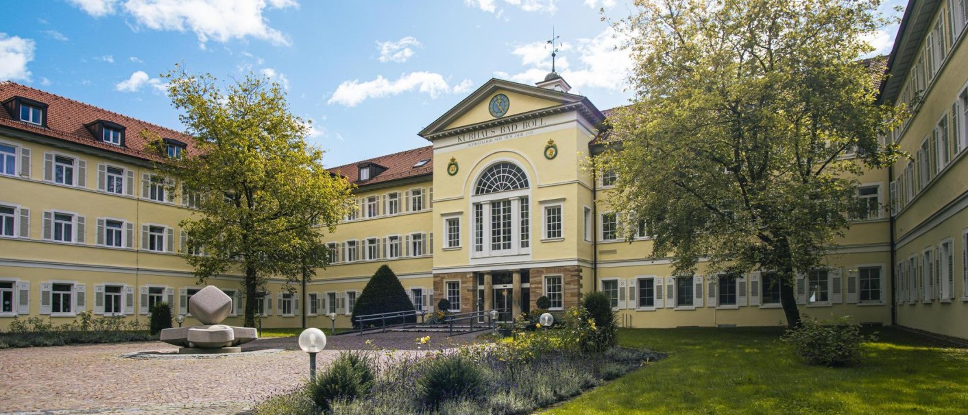 Das Kurhaus Bad Boll zeigt eine gelbe Fassade mit vielen Fenstern. Im Vordergrund sind ein Baum und ein gepflegter Garten zu sehen., &copy; SMG, Sarah Schmid