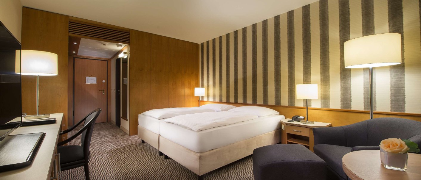 Modernes Hotelzimmer mit Doppelbett, Schreibtisch, Sessel und stilvoller Beleuchtung. Die Wände sind mit Streifenmuster tapeziert., © Maritim Hotelgesellschaft mbH