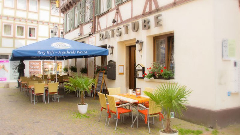 Ein Restaurant namens Ratsstube mit Außenbestuhlung unter einem blauen Sonnenschirm. Fachwerkgebäude im Hintergrund, Pflanzen schmücken den Eingangsbereich. Ein Restaurant namens Ratsstube mit Außenbestuhlung unter einem blauen Sonnenschirm. Fachwerkgebäude im Hintergrund, Pflanzen schmücken den Eingangsbereich.