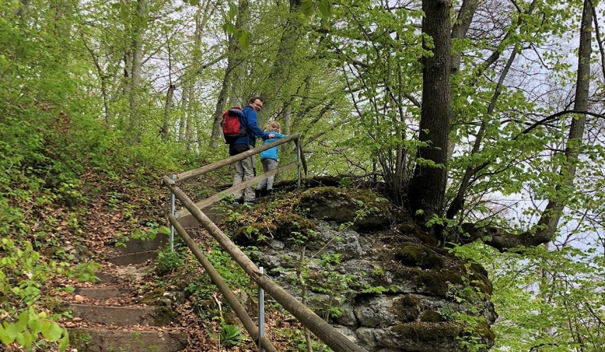 Zwei Wanderer stehen auf einem bewaldeten Pfad mit Geländer. Der Weg führt durch einen grünen Wald mit Laub und Felsen., © Touristik und Marketing GmbH