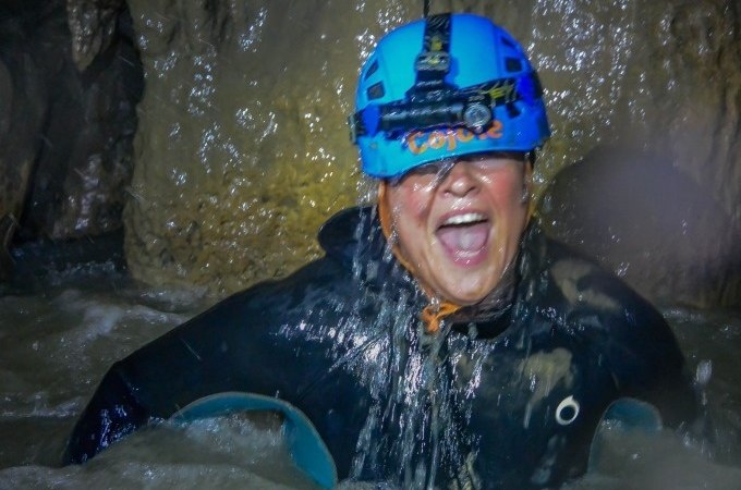 Eine Person mit blauem Helm und Neoprenanzug lacht in einer nassen Höhle. Sie ist von Wasser umgeben und wirkt abenteuerlustig., © SwabianTravel