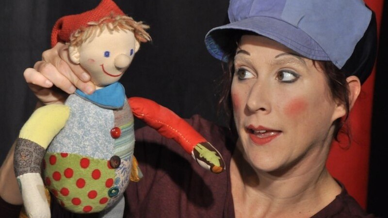 Frau mit blauer Mütze hält eine bunte Stoffpuppe. Sie hat rote Wangen und schaut die Puppe an. Hintergrund ist dunkel mit rotem Vorhang., © Mauerwerk GmbH