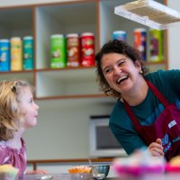 Eine Frau in einer Schürze zeigt einem Kind eine Schokoladenform. Im Hintergrund sind bunte Dosen zu sehen., © Alfred Ritter GmbH &amp; Co. KG | Waldenbuch