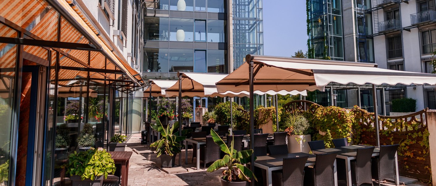 Moderne Terrasse mit Tischen, Stühlen und Pflanzen unter Sonnenschirmen vor einem Glasgebäude., © Plaza Residence Stuttgart-Ditzingen Moderne Terrasse mit Tischen, Stühlen und Pflanzen unter Sonnenschirmen vor einem Glasgebäude., © Plaza Residence Stuttgart-Ditzingen