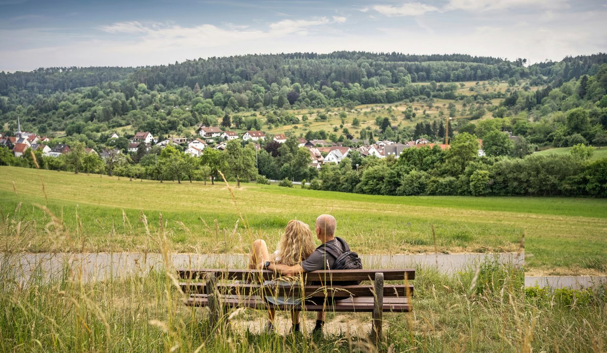 Ein Paar sitzt auf einer Bank und genießt den Ausblick auf eine grüne Landschaft mit einem Dorf im Hintergrund. Ein Paar sitzt auf einer Bank und genießt den Ausblick auf eine grüne Landschaft mit einem Dorf im Hintergrund.