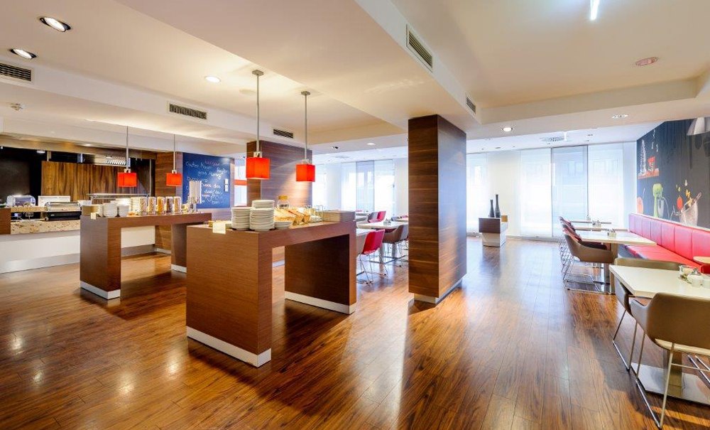 Moderner Frühstücksbereich mit Holzboden, Tischen, roten Lampen und Sitzgelegenheiten. Helle, einladende Atmosphäre mit Buffetbereich., © Ibis Stuttgart Airport Messe Moderner Frühstücksbereich mit Holzboden, Tischen, roten Lampen und Sitzgelegenheiten. Helle, einladende Atmosphäre mit Buffetbereich., © Ibis Stuttgart Airport Messe