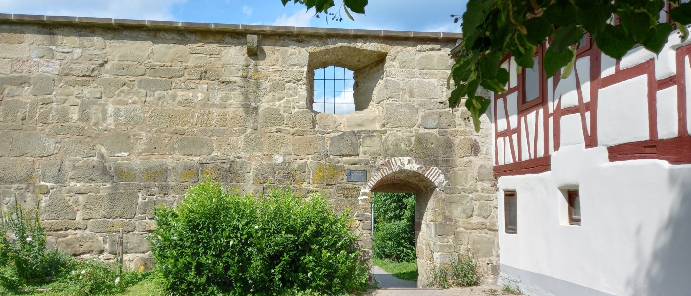 Historische Stadtmauer mit kleinem Torbogen und leicht nach links versetztem "Fenster", rechts an die Mauer anschlie&szlig;end ein Fachwerkhaus mit roten Holzbalken. Im Vordergrund eine Wiese mit Str&auml;uchern., &copy; Petra Natzkowski