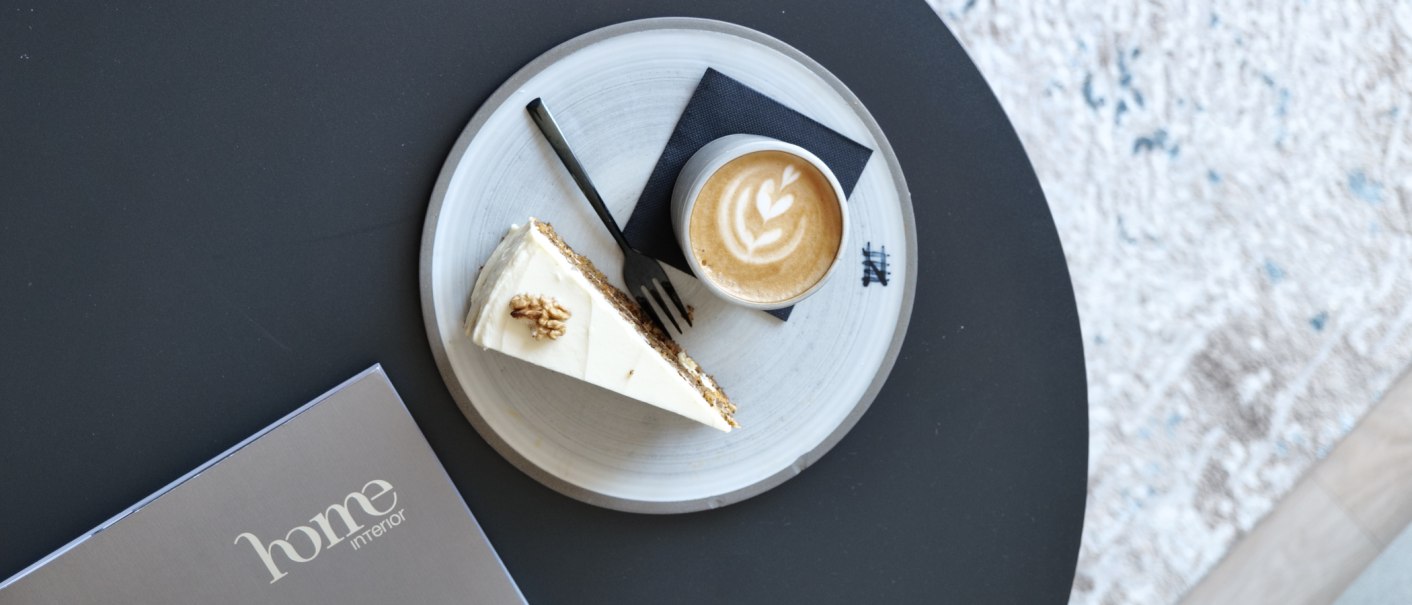 Latte mit Herz-Latte-Art und ein St&uuml;ck Kuchen auf einem Teller auf schwarzem Tisch, daneben eine Karte mit &bdquo;home interior&ldquo;., &copy; Taraba