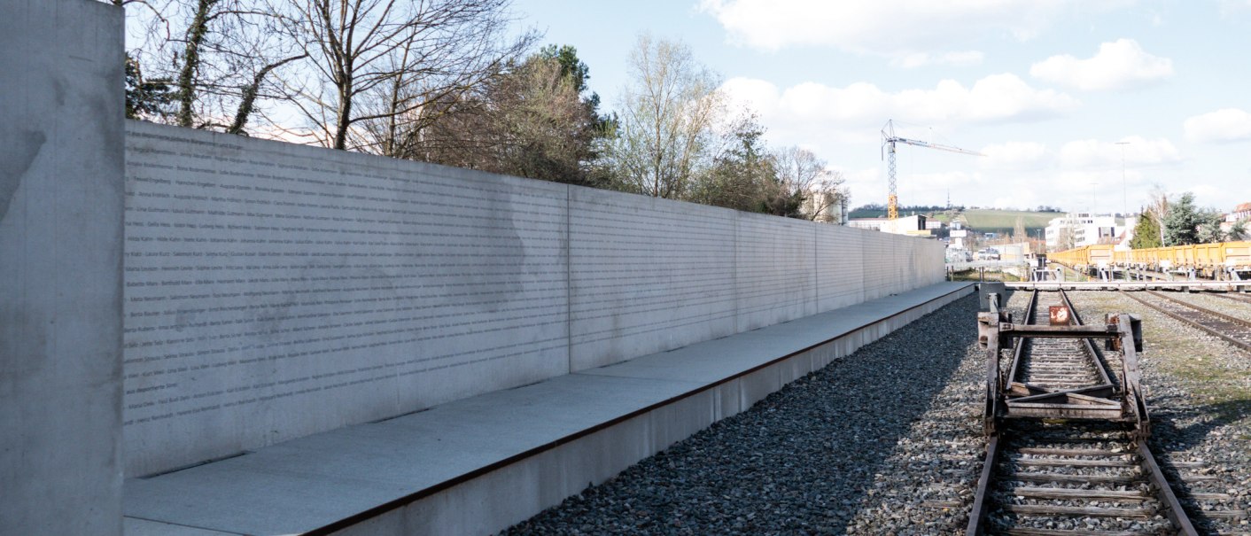 Eine lange Betonwand mit eingravierten Namen steht neben stillgelegten Bahngleisen unter einem bewölkten Himmel., © Foto-Kunst Andreas Keller