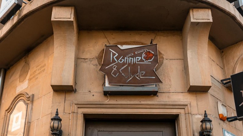 Ein Schild mit der Aufschrift 'Bonnie & Clyde' h&auml;ngt an einem Geb&auml;ude. Die Fassade ist aus Stein, und es gibt dekorative Laternen., &copy; SMG. Sarah Schmid