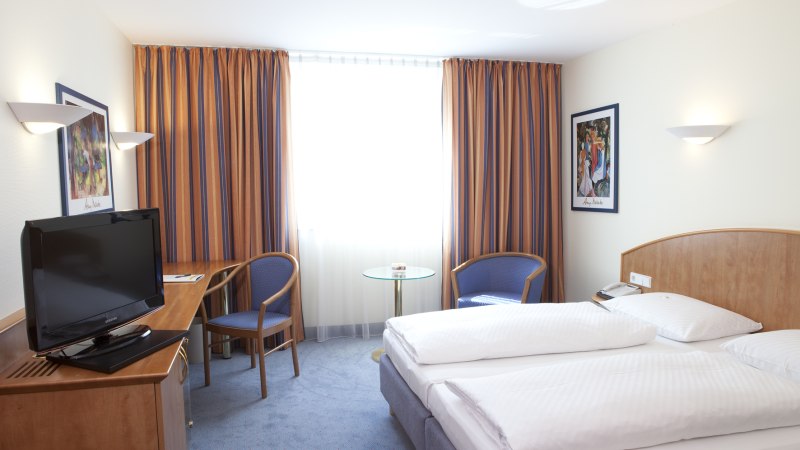 Ein Hotelzimmer mit Doppelbett, Fernseher, zwei Stühlen und einem kleinen Tisch vor einem Fenster mit gestreiften Vorhängen., © Hotel Schwanen Stuttgart-Airport/Messe