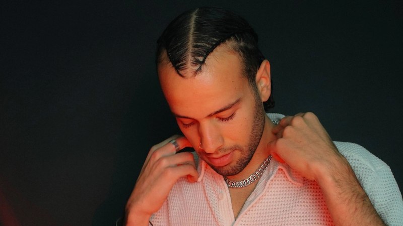 Ein Mann mit geflochtenem Haar und weißem Hemd blickt nach unten und hält seine Halskette fest., © Kulturhaus Schwanen